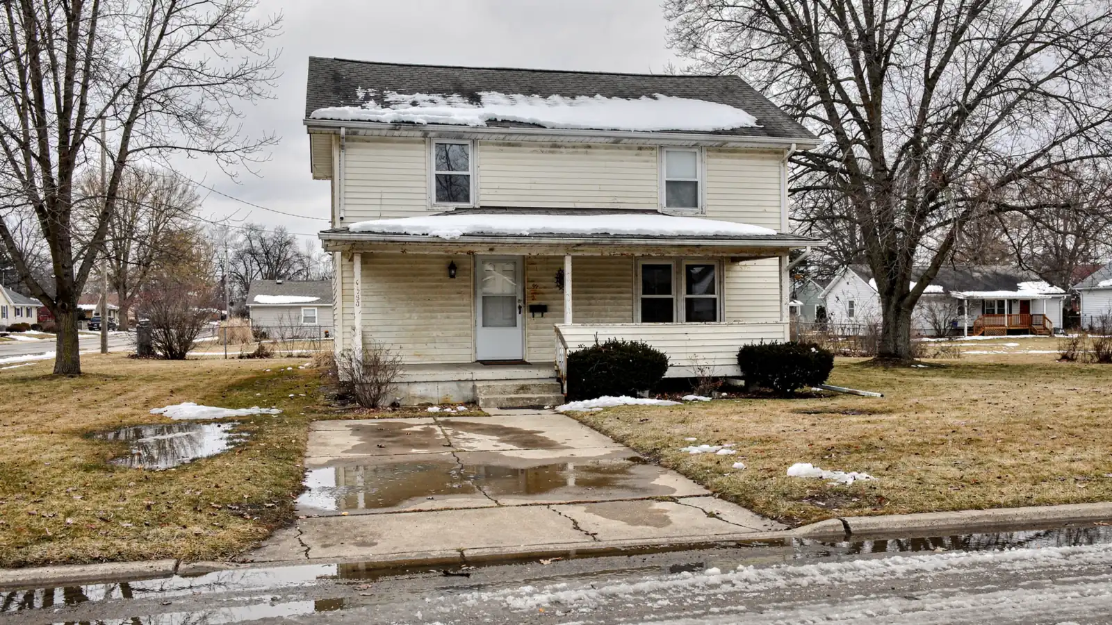 A plain clapboard house on a flat lot with standing meltwater across the lawn and driveway in late winter.